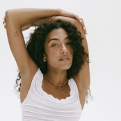 A woman with brown curly hair with a white vest on stood in front of a white background